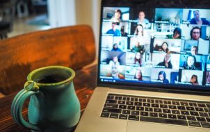 A laptop displaying a grid view of a virtual meeting with multiple participants on screen, alongside a turquoise ceramic mug on a wooden table.