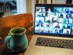 A laptop displaying a grid view of a virtual meeting with multiple participants on screen, alongside a turquoise ceramic mug on a wooden table.